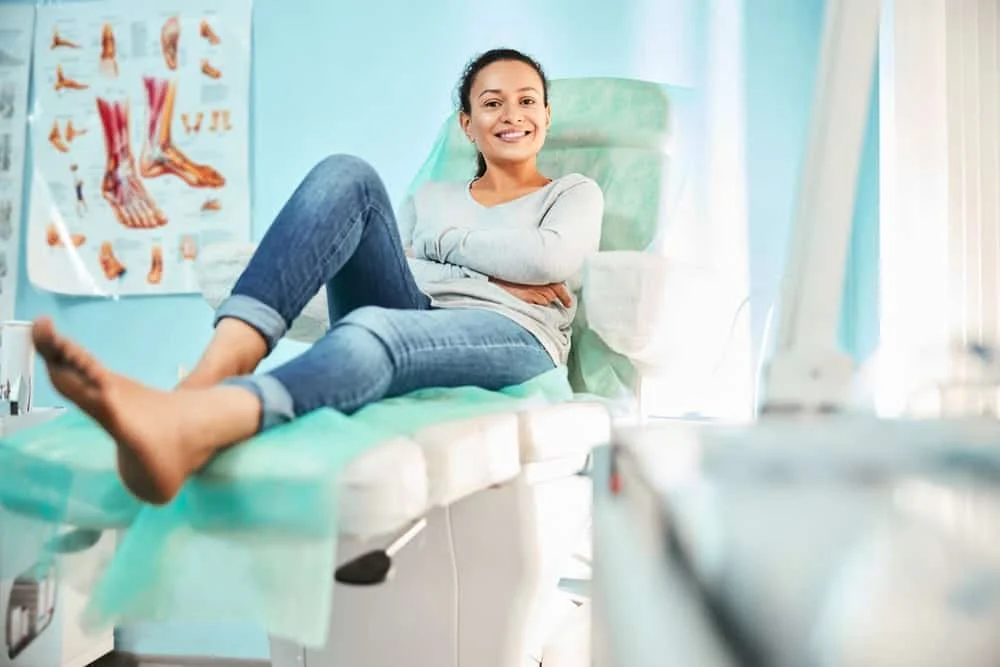 Patient relaxing barefoot in a podiatry exam chair beside a foot anatomy chart